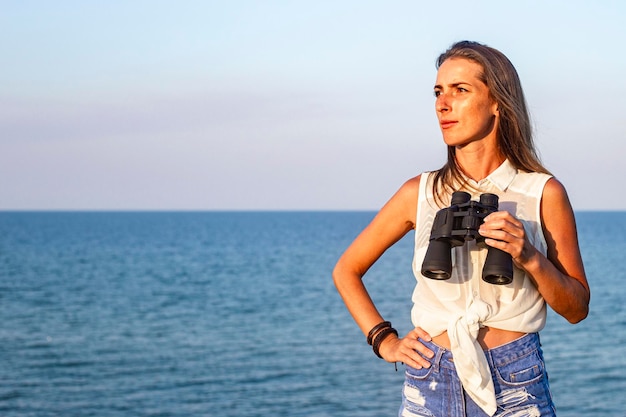 Mulher jovem e bonita com binóculos na falésia olha para o mar.