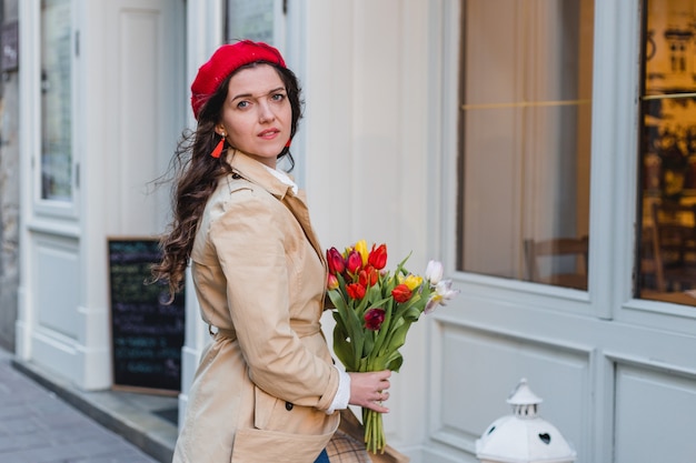 Mulher jovem e bonita com buquê de flores de tulipas da primavera na rua da cidade. Garota feliz caminhando ao ar livre. Retrato de primavera de mulher bonita na cidade velha