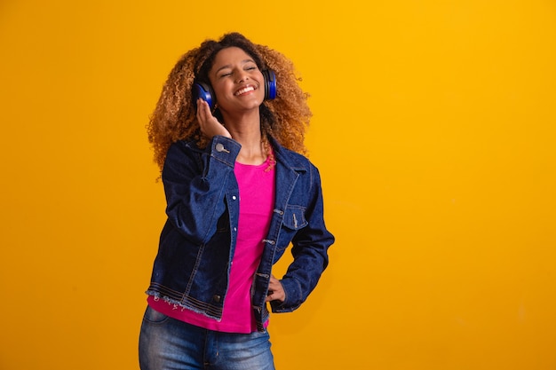 Mulher jovem e bonita com cabelo afro, ouvindo música com seu fone de ouvido em fundo amarelo, com espaço livre para texto.
