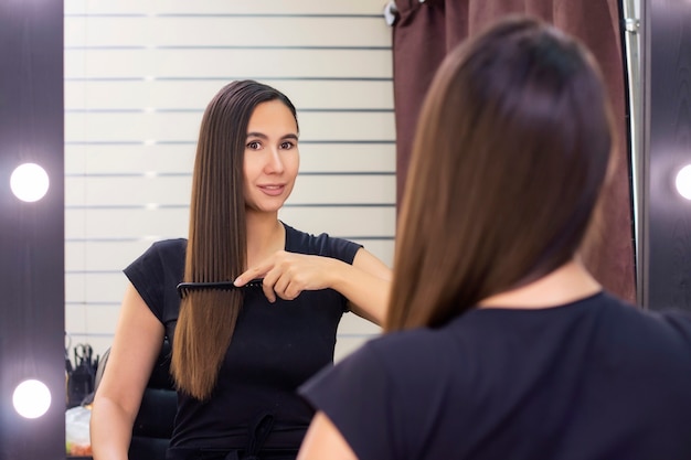 Mulher jovem e bonita com cabelo castanho comprido e liso cuida do cabelo. penteando o cabelo e se olhando no espelho