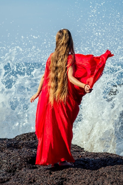 Mulher jovem e bonita com cabelo comprido, em um elegante vestido vermelho longo chique de luxo na praia com ondas. conceito de férias chiques em um resort tropical