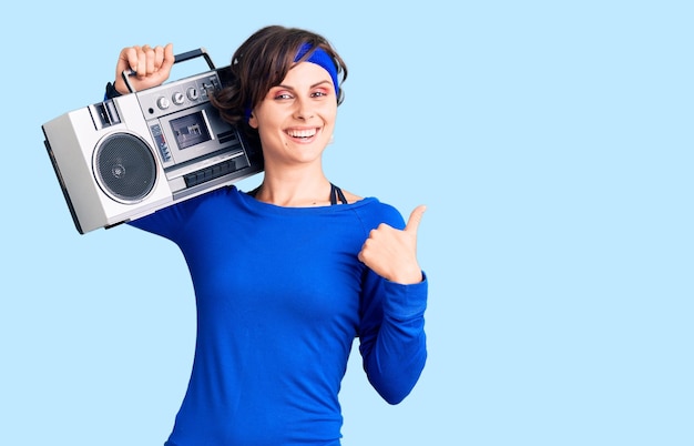 Mulher jovem e bonita com cabelo curto vestindo roupas de ginástica e segurando boombox sorrindo feliz e positivo polegar para cima fazendo excelente e sinal de aprovação