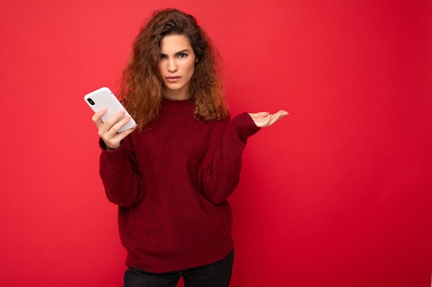 Mulher jovem e bonita com cabelo encaracolado, vestindo um suéter vermelho escuro isolado em um fundo vermelho segurando o celular