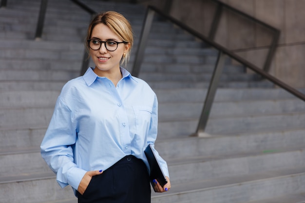 Mulher jovem e bonita com cabelo loiro em pé na escada e segurando o tablet digital. Mulher sorridente usando óculos, camisa azul e calça preta.