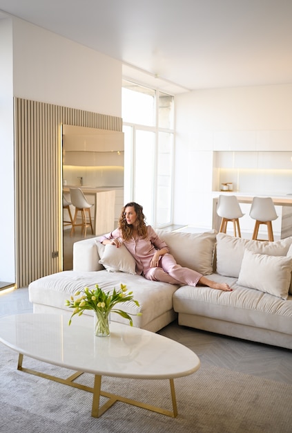 Mulher jovem e bonita com cabelos cacheados de pijama rosa deitado no sofá do sofá branco de manhã. sala de estar de estilo escandinavo e interior da cozinha.