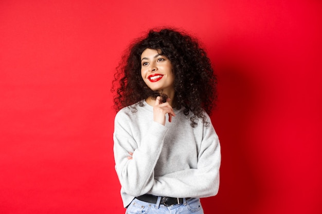 Mulher jovem e bonita com cabelos cacheados e lábios vermelhos, brincando com cachos e olhando para o lado, olhando romanticamente, de pé no fundo do estúdio.