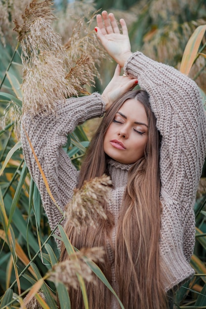 Mulher jovem e bonita com cabelos compridos e olhos castanhos no parque outono Retrato de um modelo em um suéter de malha perto da grama dos pampas Queda