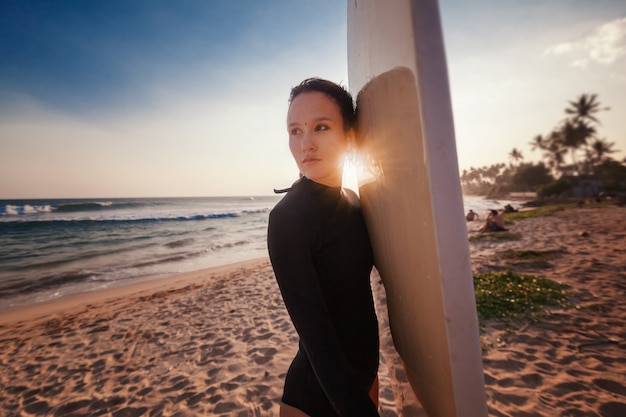 Foto mulher jovem e bonita com cabelos escuros com uma prancha de surf ao sol pôr do sol no oceano