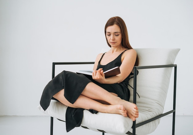 Mulher jovem e bonita com cabelos longos escuros em vestido preto lendo livro na cadeira no interior branco