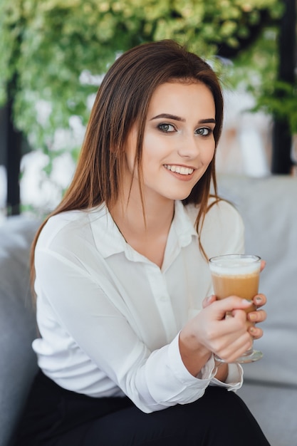 Mulher jovem e bonita com café nas mãos em uma camisa branca, sentado no escritório. fundo desfocado.