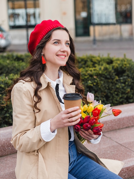 Mulher jovem e bonita com café para viagem e buquê de flores de tulipas da primavera na rua da cidade.