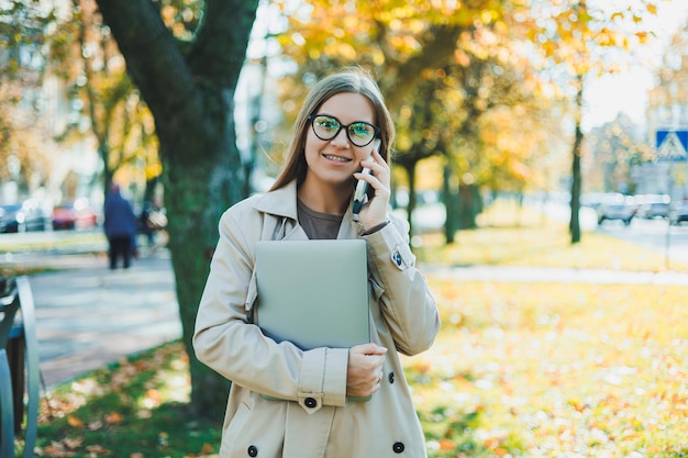 Mulher jovem e bonita com casaco de outono usando laptop e telefone trabalhando remotamente mulher falando no celular no parque de outono