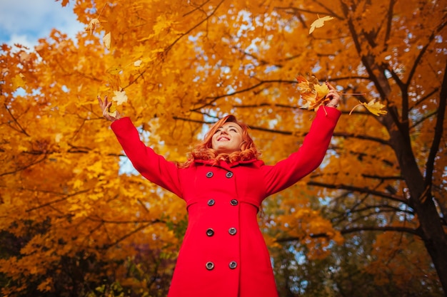 Foto mulher jovem e bonita com casaco vermelho jogando folhas na floresta de outono