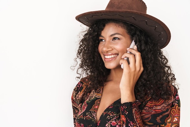 Mulher jovem e bonita com chapéu de cowboy em pé, isolado na parede branca, falando no celular