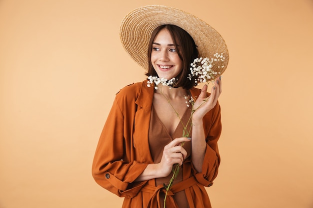 Foto mulher jovem e bonita com chapéu de palha em pé, isolado sobre uma parede bege, posando com flores