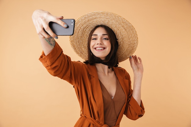 Mulher jovem e bonita com chapéu de palha isolado sobre uma parede bege, tirando uma selfie
