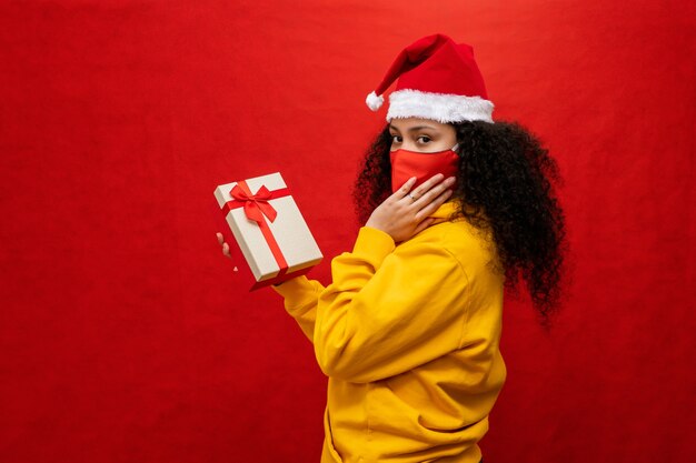 Foto mulher jovem e bonita com chapéu de papai noel e máscara protetora de coronavirus, ela está segurando caixas de presente.