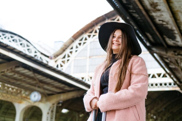 Mulher jovem e bonita com chapéu preto e casaco rosa com mala fica na plataforma da estação tou ...