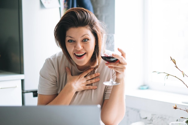 Mulher jovem e bonita com corpo positivo com copo de vinho tendo chamada de vídeo no laptop