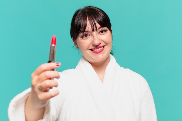 Mulher jovem e bonita com expressão feliz de roupão de banho e segurando um batom