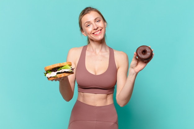 Mulher jovem e bonita com expressão feliz segurando um sanduíche e uma rosquinha