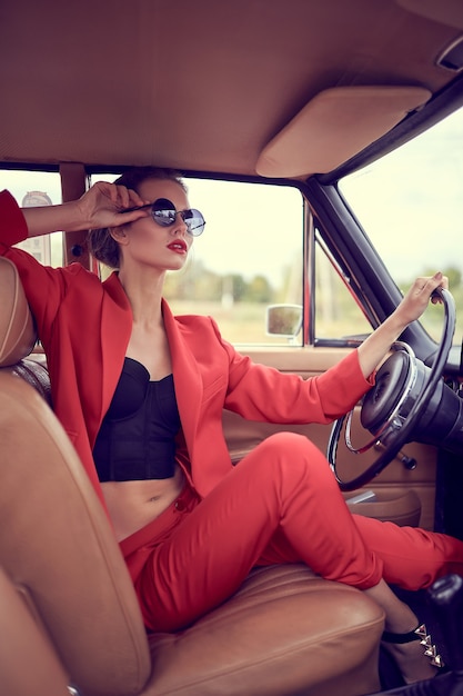 Mulher jovem e bonita com fantasia vermelha e óculos escuros sentada em um carro retrô