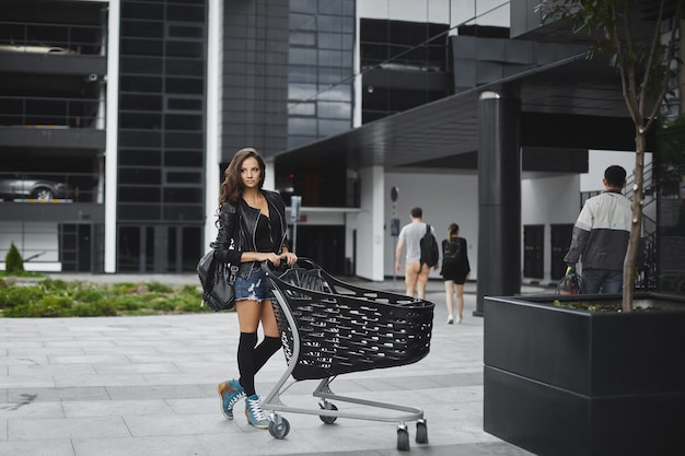 Mulher jovem e bonita com longos cabelos escuros em shorts jeans e jaqueta de couro, posando com um carrinho de compras perto do exterior da loja.