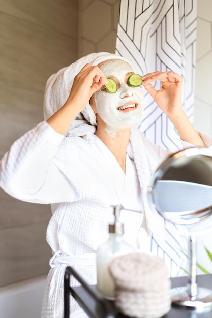 Mulher jovem e bonita com máscara facial no rosto, segurando fatias de pepino fresco.