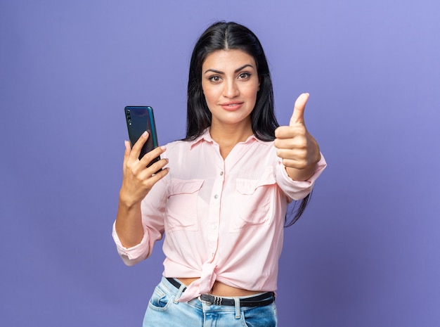 Mulher jovem e bonita com roupas casuais segurando um smartphone, parecendo feliz e confiante, mostrando os polegares para cima sorrindo