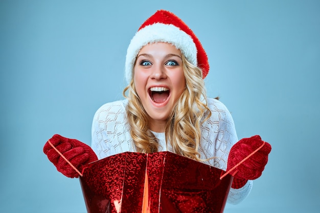 Mulher jovem e bonita com roupas de Papai Noel com um presente em um fundo azul