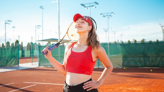 Mulher jovem e bonita com roupas esportivas vermelhas, posando com uma raquete de tênis na quadra