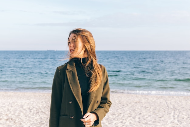 Mulher jovem e bonita com um casaco verde, caminhando ao longo da costa