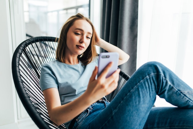 Mulher jovem e bonita com um celular sentada em uma cadeira perto da janela em casa