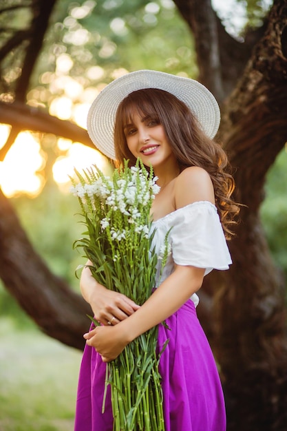 Mulher jovem e bonita com um chapéu branco com um buquê de flores silvestres ao pôr do sol no parque