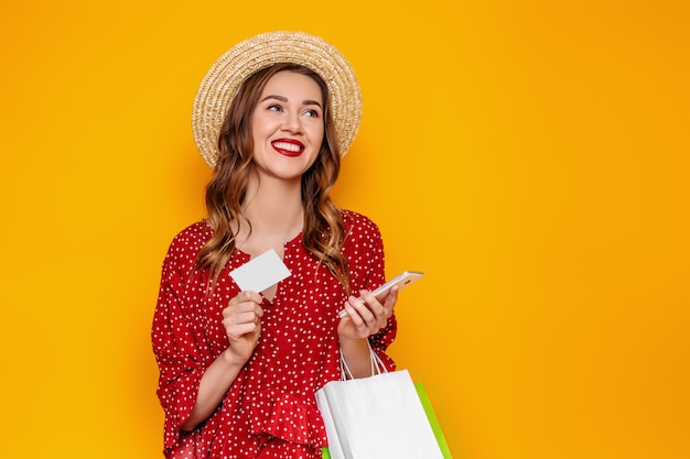 Mulher jovem e bonita com um chapéu de palha vermelho vestido de verão possui um telefone celular e um cartão de crédito nas mãos dela isolado em um banner de web de maquete de parede amarelo. Garota faz compras on-line