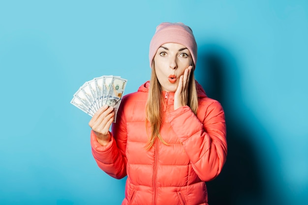 Mulher jovem e bonita com um chapéu e uma jaqueta de inverno com uma cara surpresa segurando dinheiro