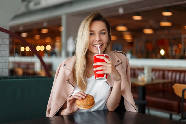 Mulher jovem e bonita com um hambúrguer bebe refrigerante em um café