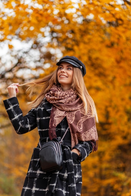 Mulher jovem e bonita com um sorriso em um casaco da moda com um lenço e um chapéu caminha em um parque de outono com folhagem amarela