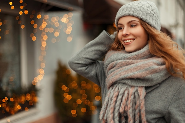 Mulher jovem e bonita com um sorriso magnífico em um casaco cinza da moda e um chapéu de malha de inverno com um lenço vintage, caminhando ao ar livre nas férias