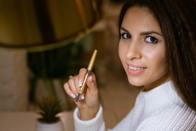 Mulher jovem e bonita com um suéter branco e uma caneta na mão sentada pensativa à mesa