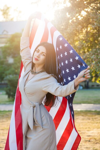 Mulher jovem e bonita com um vestido clássico segurando a bandeira americana no parque.