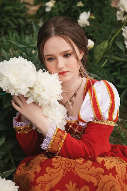 Mulher jovem e bonita com um vestido vermelho medieval segurando peônias brancas