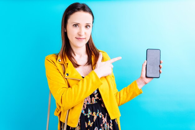 Mulher jovem e bonita com uma jaqueta amarela sobre fundo azul apontando e mostrando um smartphone