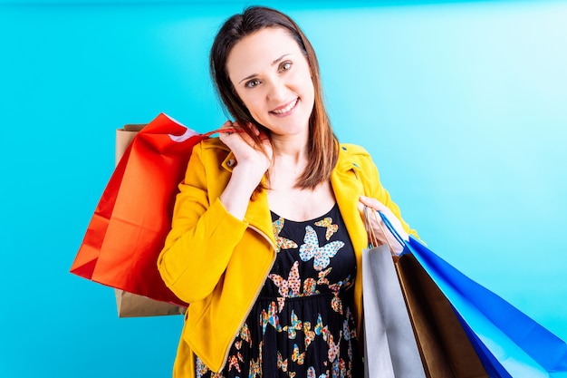 Mulher jovem e bonita com uma jaqueta amarela sobre fundo azul. feliz com sacolas de compras