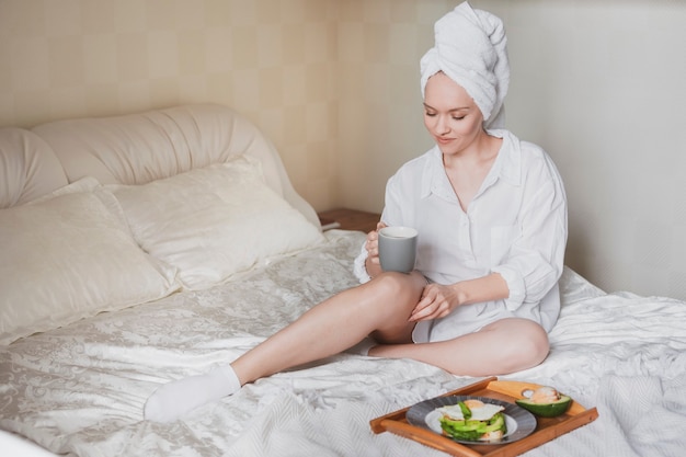 Mulher jovem e bonita com uma toalha na cabeça na cama tomando um café da manhã saudável