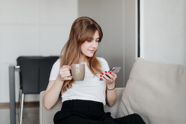 Mulher jovem e bonita com uma xícara de café, olhando para o telefone, sentado no sofá em casa, no apartamento
