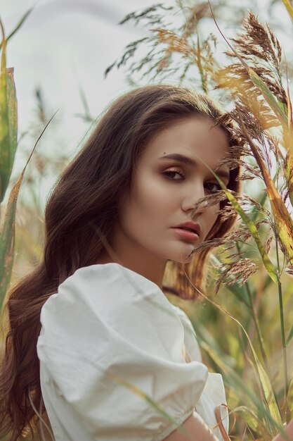 Mulher jovem e bonita com vestido branco de verão está sentada na grama alta no campo rural retrato de uma garota romântica ao pôr do sol sol quente mulher de beleza natural