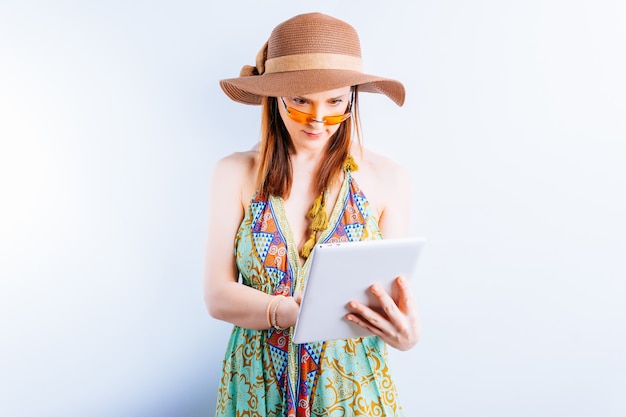 Mulher jovem e bonita com vestido de verão, chapéu e óculos amarelos sobre fundo branco com espaço de cópia, verificando o tablet do trabalho. conceito levar trabalho nas férias