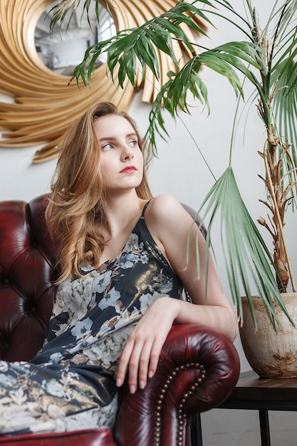 Mulher jovem e bonita com vestido heterogéneo, retrato de dentro de casa do bonito modelo pensativo. sentado na poltrona na sala aconchegante. senhora bonita natural posando em casa