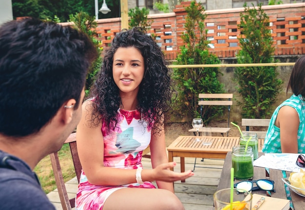 Mulher jovem e bonita conversando com um amigo ao redor da mesa com bebidas saudáveis em um dia de lazer de verão ao ar livre